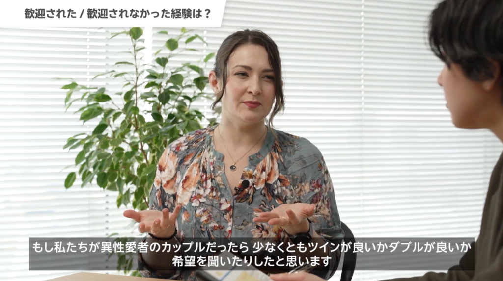 Rakuten Travel Conference 2025の従業員座談会の様子。登壇者がしゃべっており、「もし私たちが異性愛者のカップルだったら少なくともツインが良いかダブルが良いか希望を聞いたりしたと思います」という字幕が画面下部に表示されている